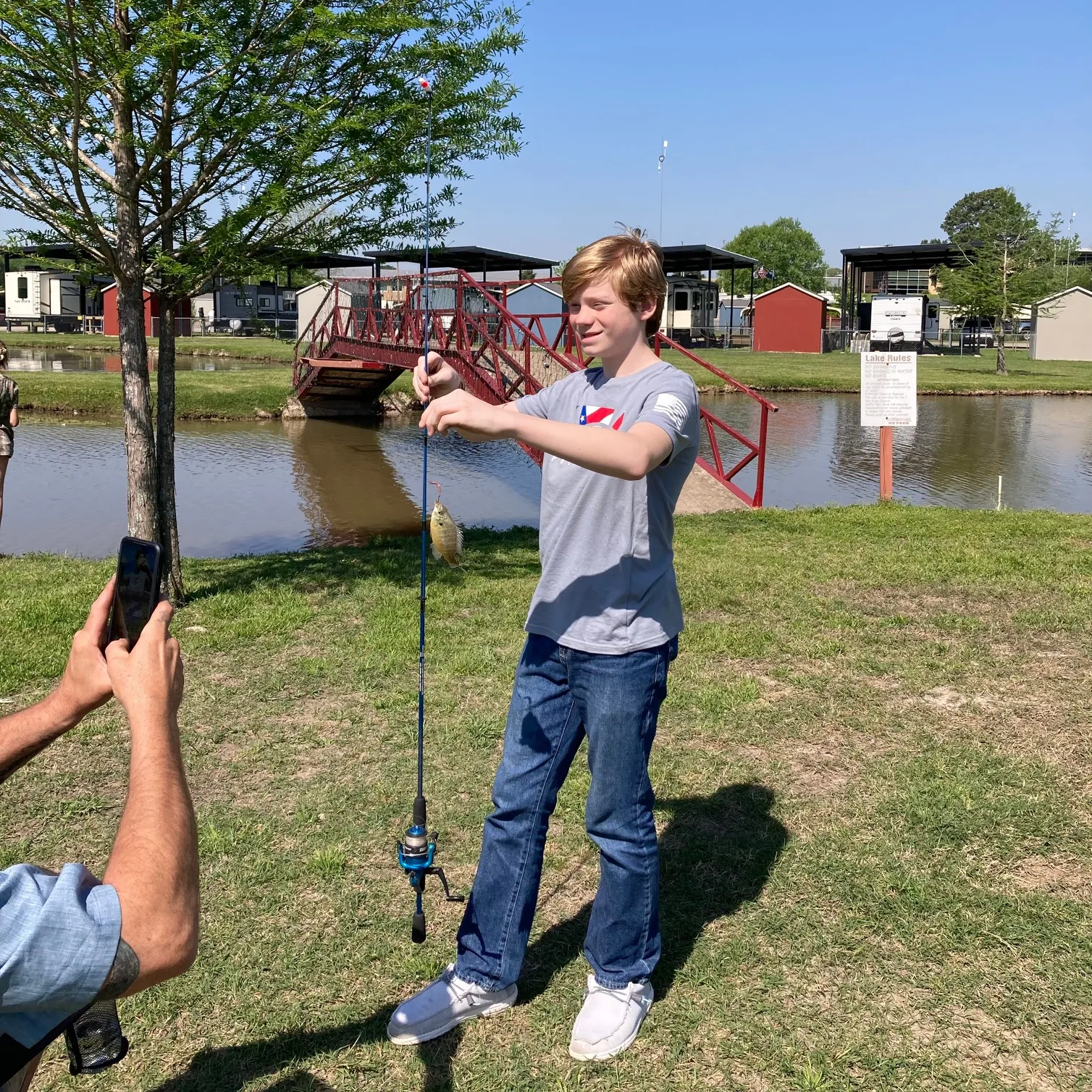 A young guest holding up a fish he caught at the private pond at Antique Capital RV Park, with the red footbridge behind him