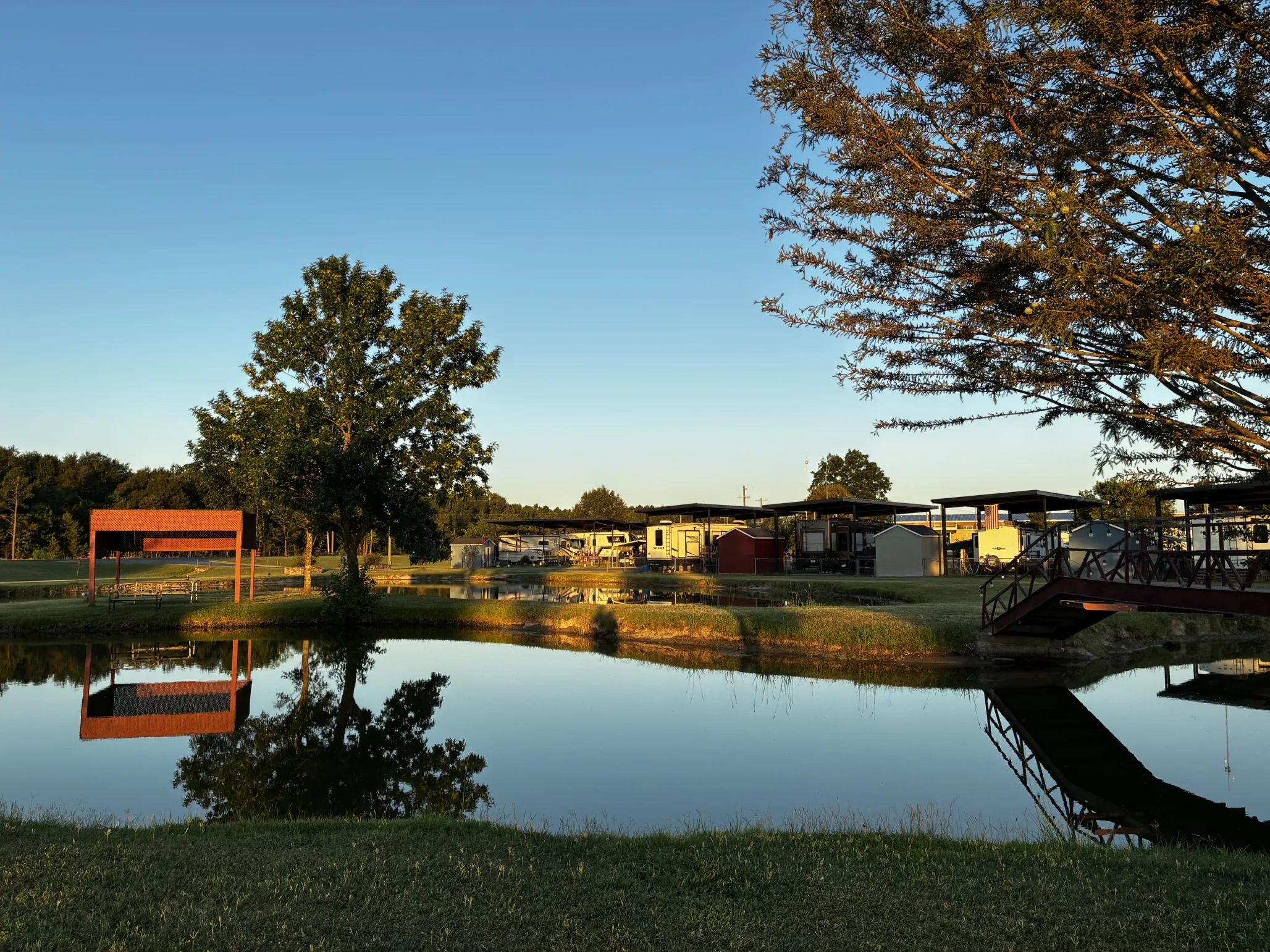 The private fishing pond and red footbridge at Antique Capital RV Park, with grassy lawns and covered RV sites in the evening light
