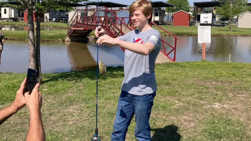 A young guest holding up a fish he caught at the private pond at Antique Capital RV Park, with the red footbridge behind him