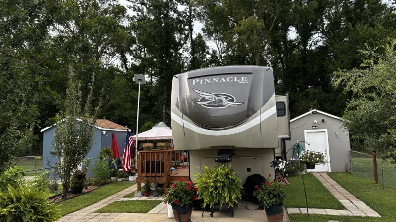 A fifth-wheel RV parked on a full-hookup site at Antique Capital RV Park with a wooden deck, potted flowers, and a paved walkway